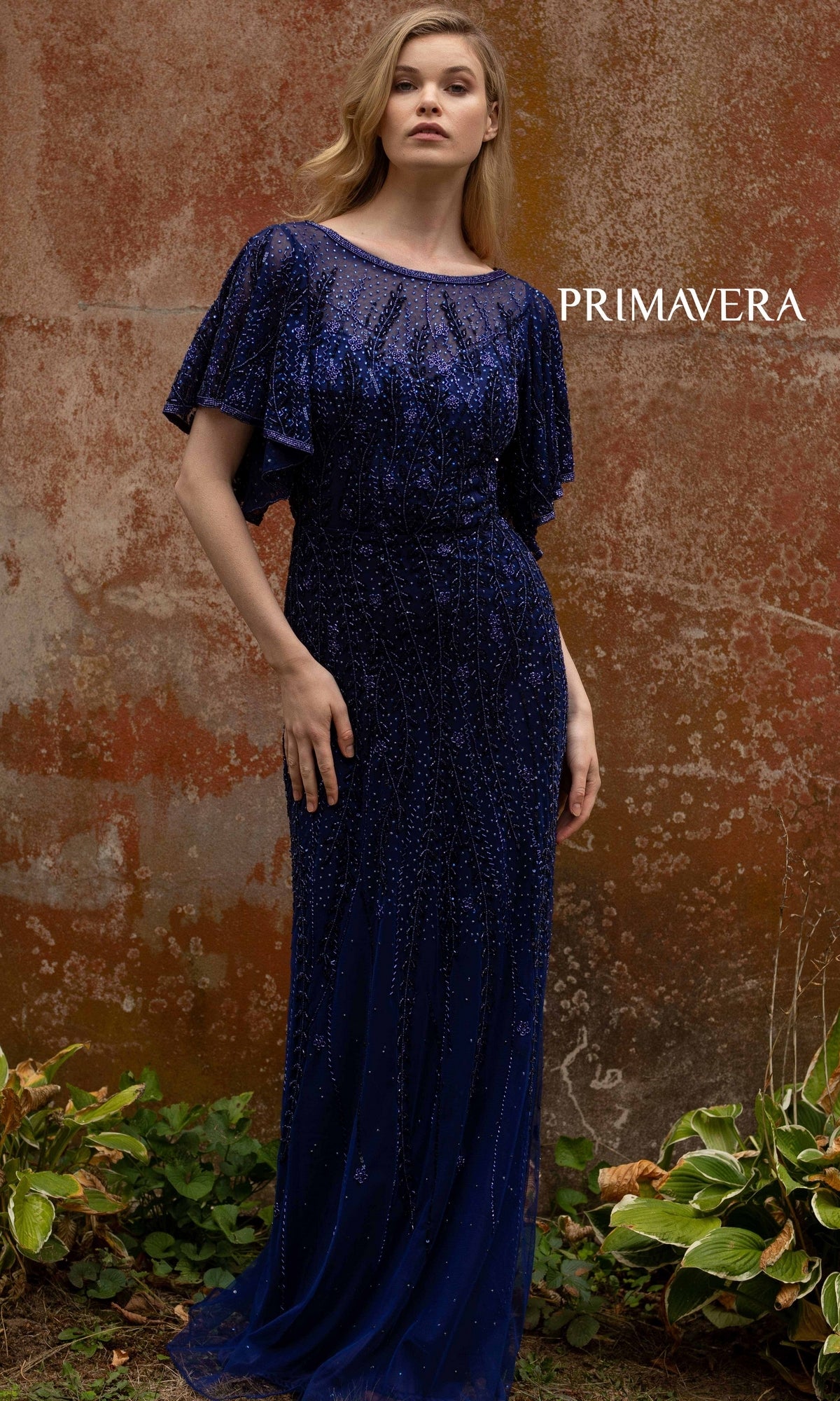 A woman stands outdoors in front of a textured wall, wearing the Short-Sleeve Beaded Long Prom Dress 3681 in dark blue. The word